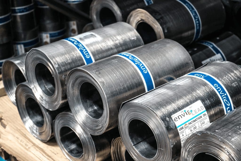 Coiled rolls of industrial metal strip stacked on a wooden pallet in a warehouse or manufacturing environment. The metallic coils are banded with blue labels and arranged in multiple layers, with additional metal stock visible in the background.