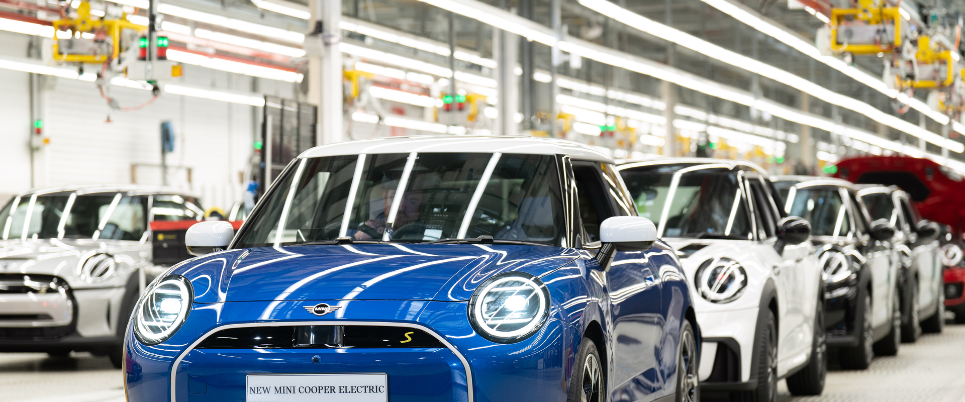 Blue MINI Cooper Electric vehicle on an automotive production line, surrounded by other MINI cars inside a modern manufacturing facility.