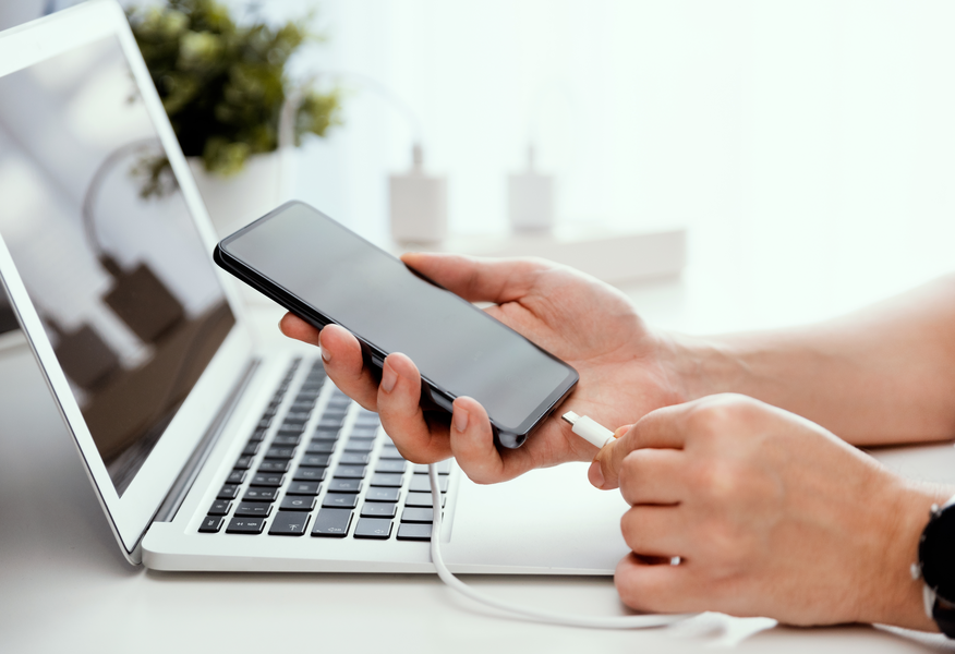 Person holding a smartphone and connecting a charging cable beside an open laptop on a desk, illustrating device charging and connectivity.