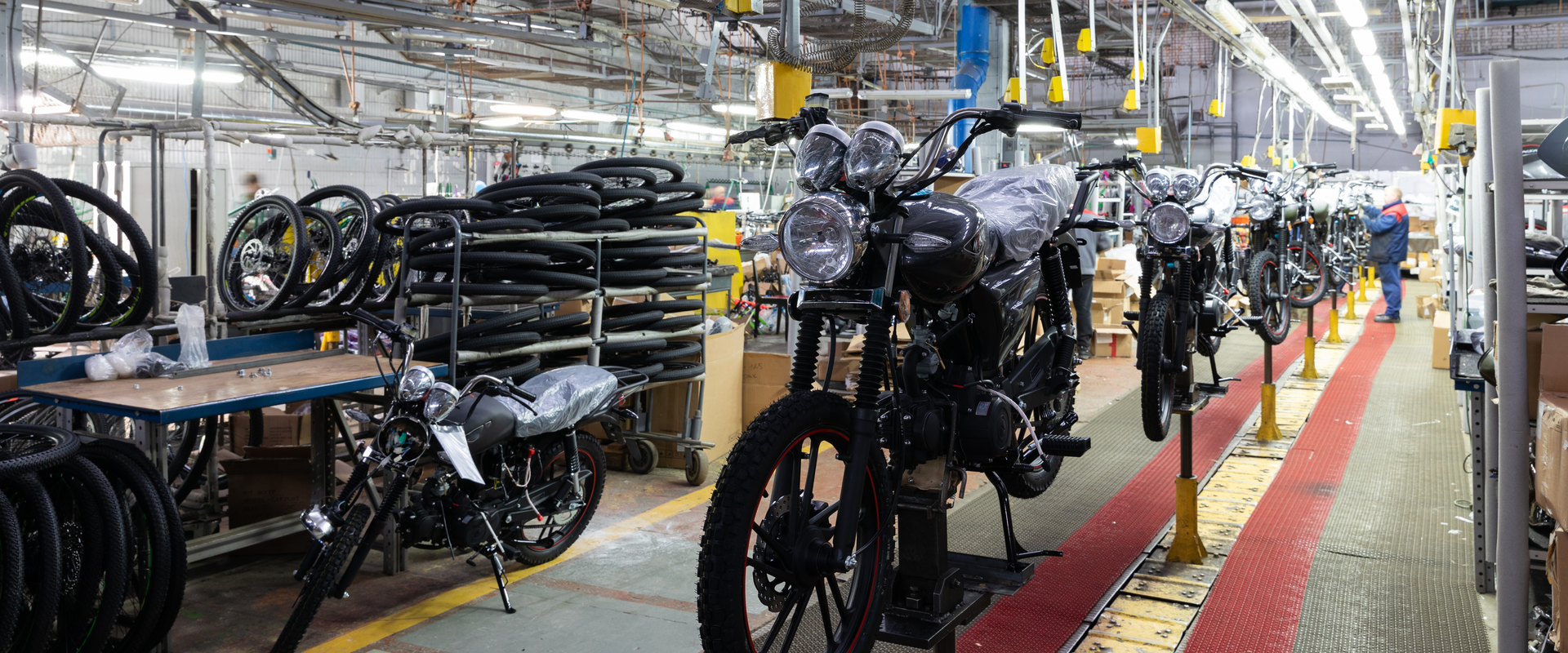 Motorcycles move along an assembly line inside a busy manufacturing facility. Several partially assembled motorcycles are positioned on production stands, with stacks of tyres, workstations, equipment, and workers visible throughout the brightly lit factory.