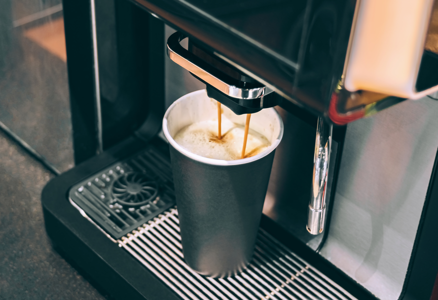 Automatic coffee machine dispensing freshly brewed coffee into a black takeaway cup, with a layer of crema forming on top.