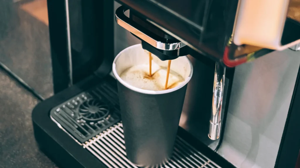 A close-up captures a coffee machine dispensing a stream of espresso into a tall black paper cup with a white interior, resting on the machine's metal drip grid. The focus is on the brewing process.