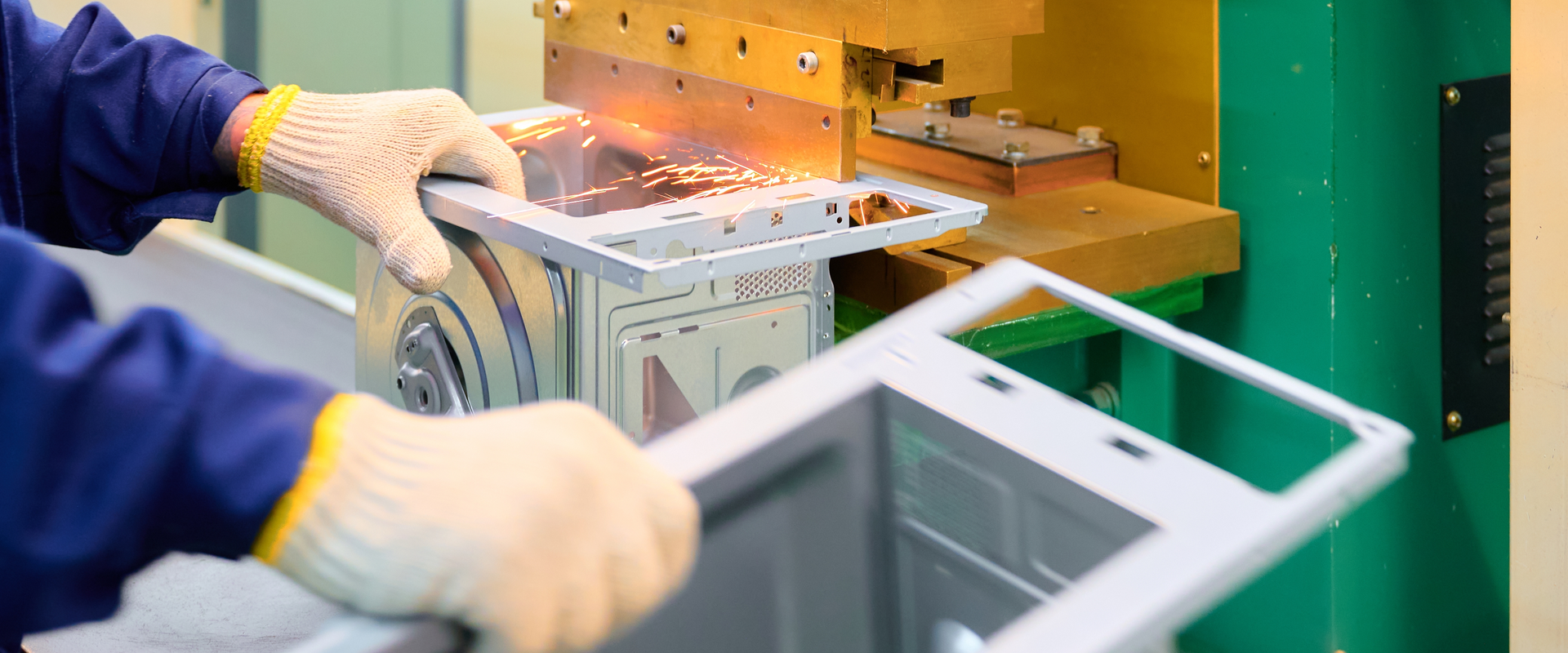 Worker wearing protective gloves positions a metal appliance housing under a manufacturing press, with sparks visible as the machine processes the component on a production line.