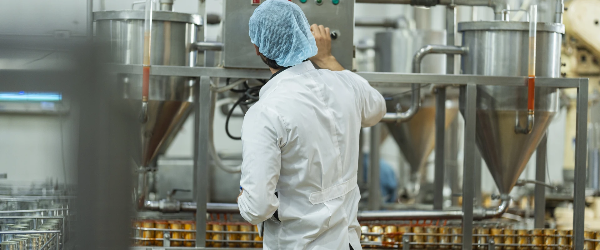 Food production facility worker wearing a white lab coat and blue hair covering operating industrial processing equipment. Large stainless steel tanks, pipes and manufacturing machinery are visible in a clean food manufacturing environment.