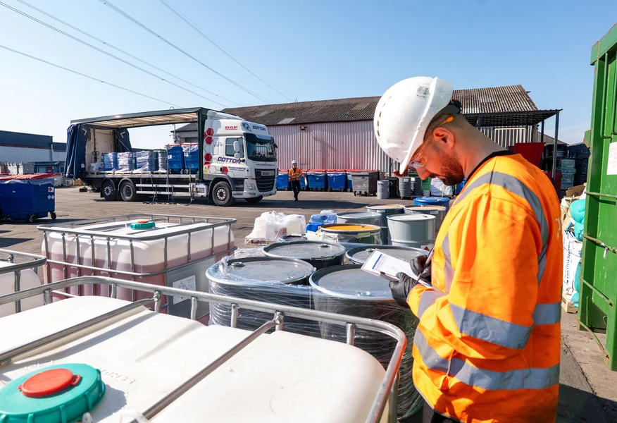 A worker wearing a white hard hat, high-visibility orange jacket, and gloves checks paperwork while standing beside large industrial containers and drums in an outdoor waste management or recycling yard. In the background, a lorry is being unloaded near a warehouse, with additional bins, containers, and equipment visible under a clear blue sky.