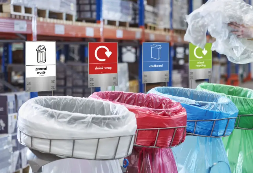 Four colour-coded recycling bins in a warehouse or industrial setting, labelled for different waste streams: general waste, shrink wrap, cardboard, and mixed recycling. A person is placing clear plastic material into the recycling area, with storage racking and inventory shelves blurred in the background.