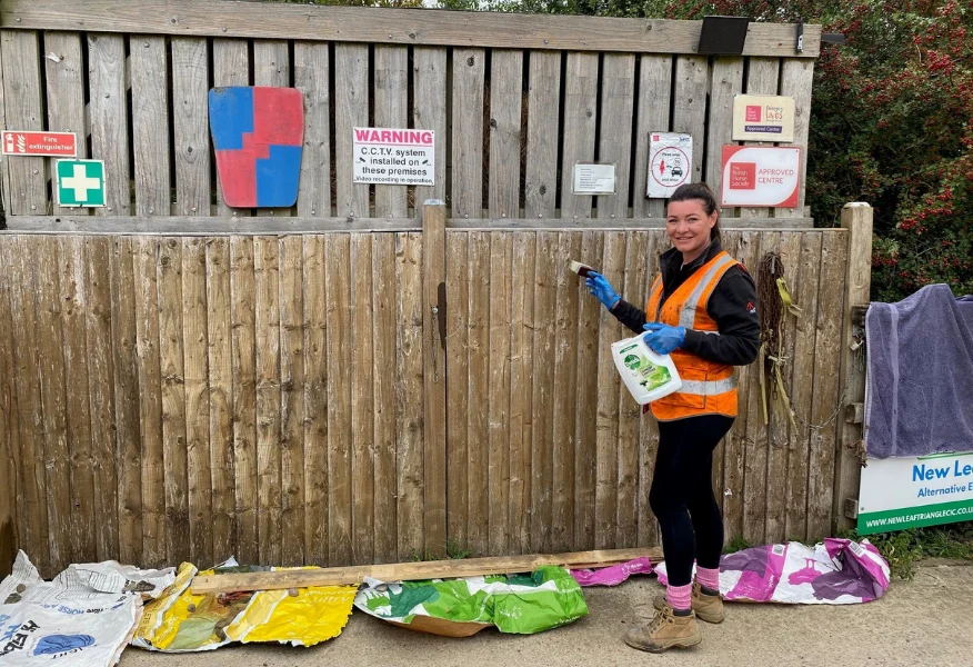 Person wearing a high-visibility orange vest, blue gloves and work boots cleans a wooden fence with a brush while holding a spray bottle. Torn bags and litter are spread on the ground in front of the fence. Several safety and accreditation signs are attached to the fence, with shrubs and trees visible in the background.