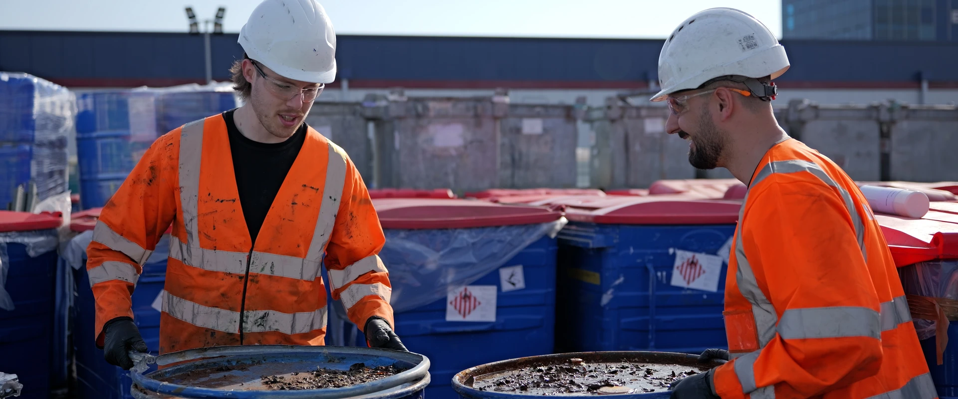 Two workers wearing high-visibility PPE and hard hats handling drums of hazardous waste at an industrial waste management facility.