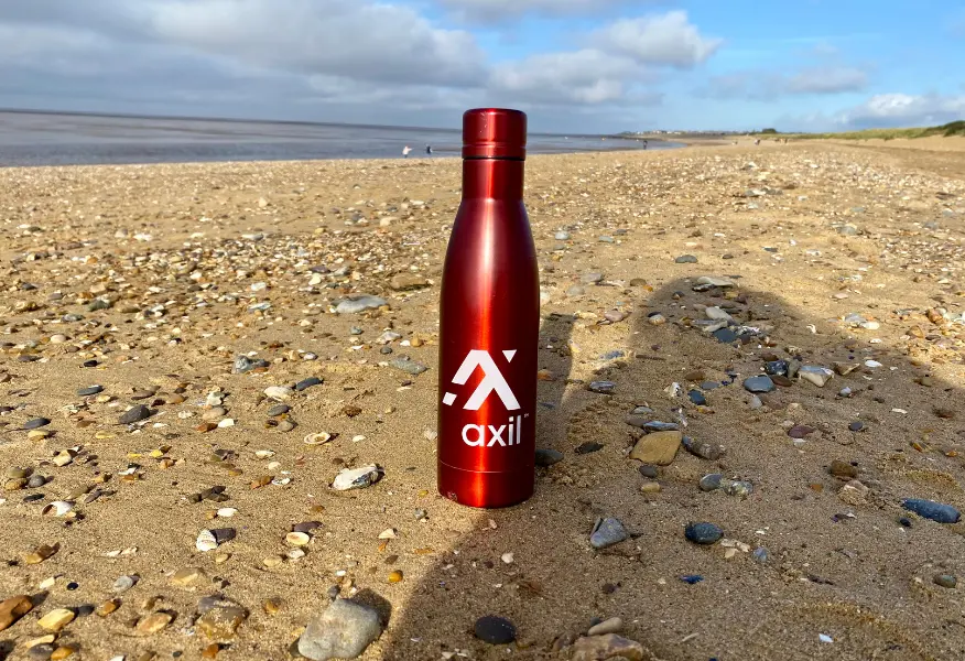 Red Axil branded reusable water bottle standing upright on a sandy, pebble-covered beach, with the sea and cloudy blue sky in the background.