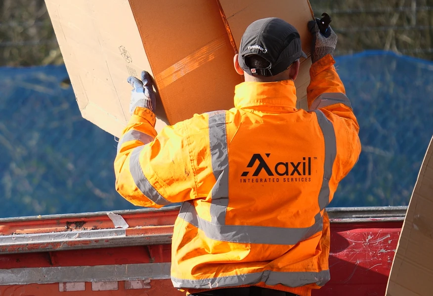 Worker in a high-visibility orange Axil Integrated Services jacket lifting a large cardboard box into a skip at a waste management or recycling site.
