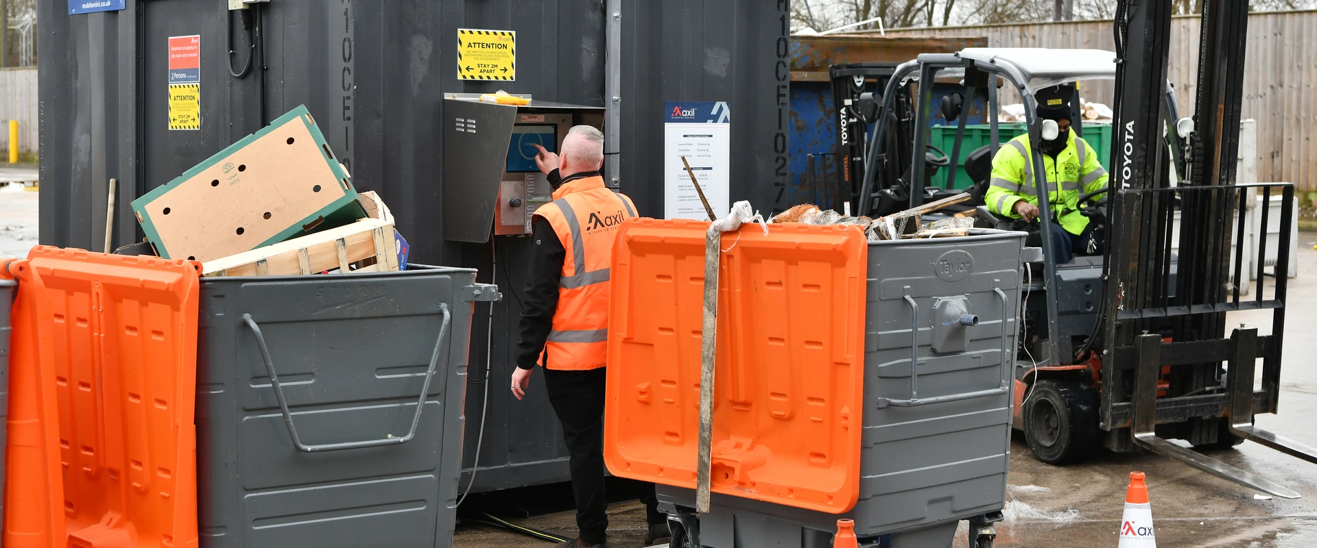 Site operative using a waste management unit while a forklift operator moves containers in a controlled work area.